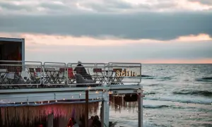 Widok na plażowy bar z widokiem na morze o zachodzie słońca, z ludźmi siedzącymi przy stolikach.