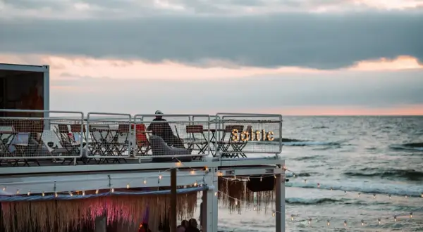 Widok na plażowy bar z widokiem na morze o zachodzie słońca, z ludźmi siedzącymi przy stolikach.